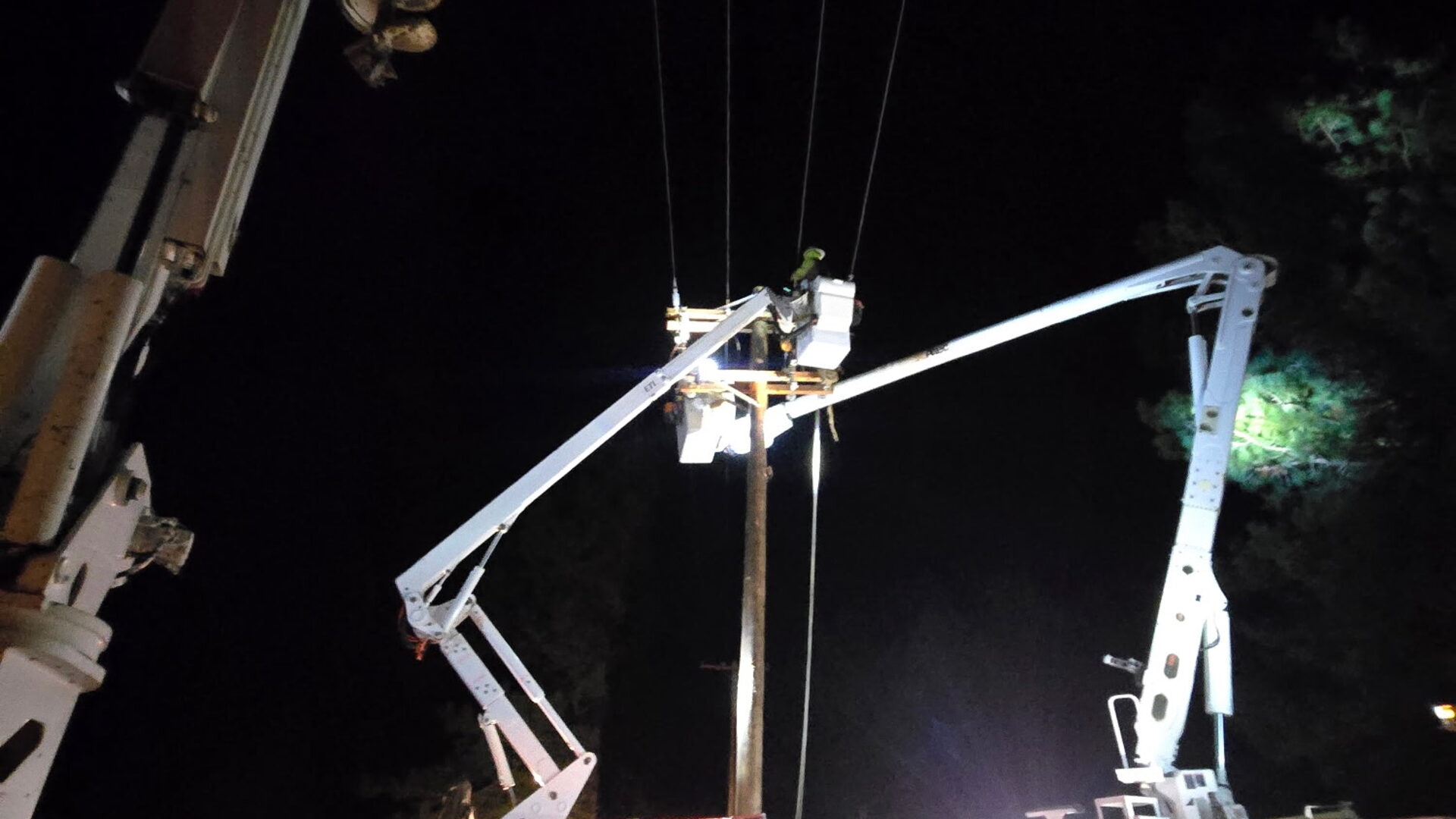 Crew performing night power-line work with floodlit bucket trucks at a wood pole