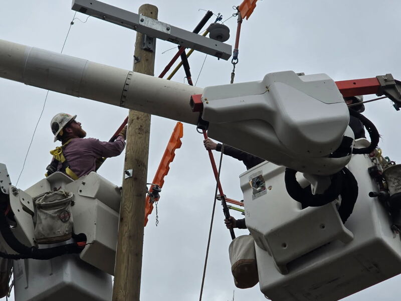 Two linemen in bucket trucks using insulated hot sticks on a wood pole with orange protective line covers