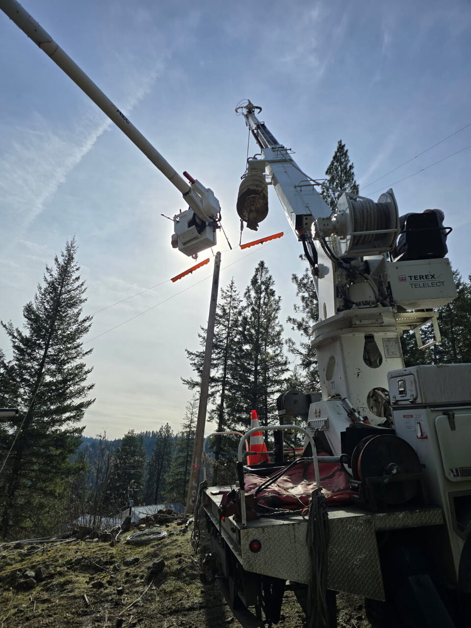 Digger derrick and bucket boom installing a wood utility pole on a wooded hillside