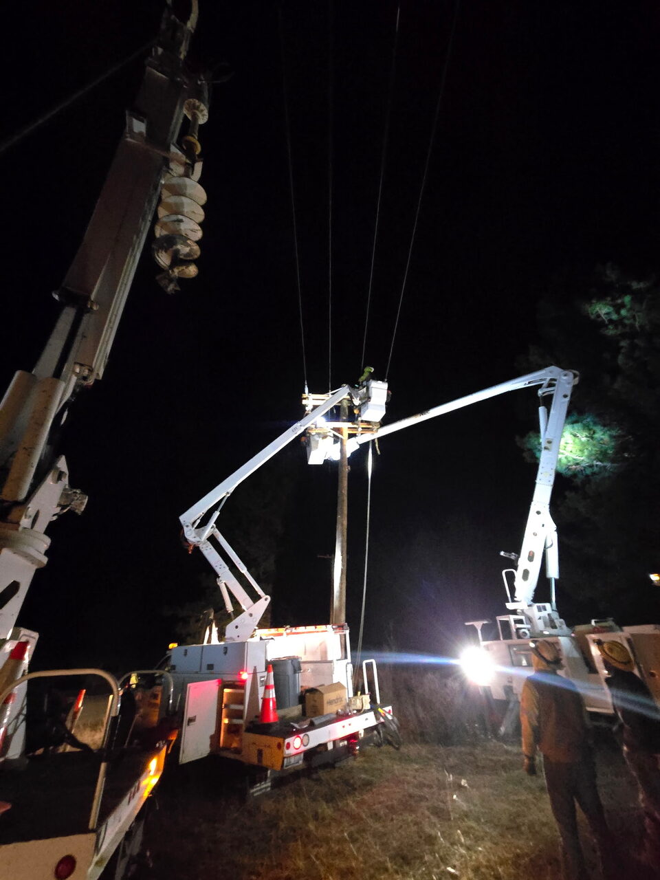 Crew performing night power-line work with floodlit bucket trucks at a wood pole